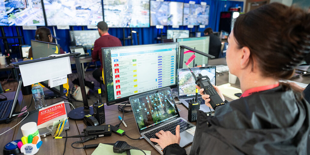 Woman holding 2-way radio viewing computer monitor in a room with a wall of monitors.